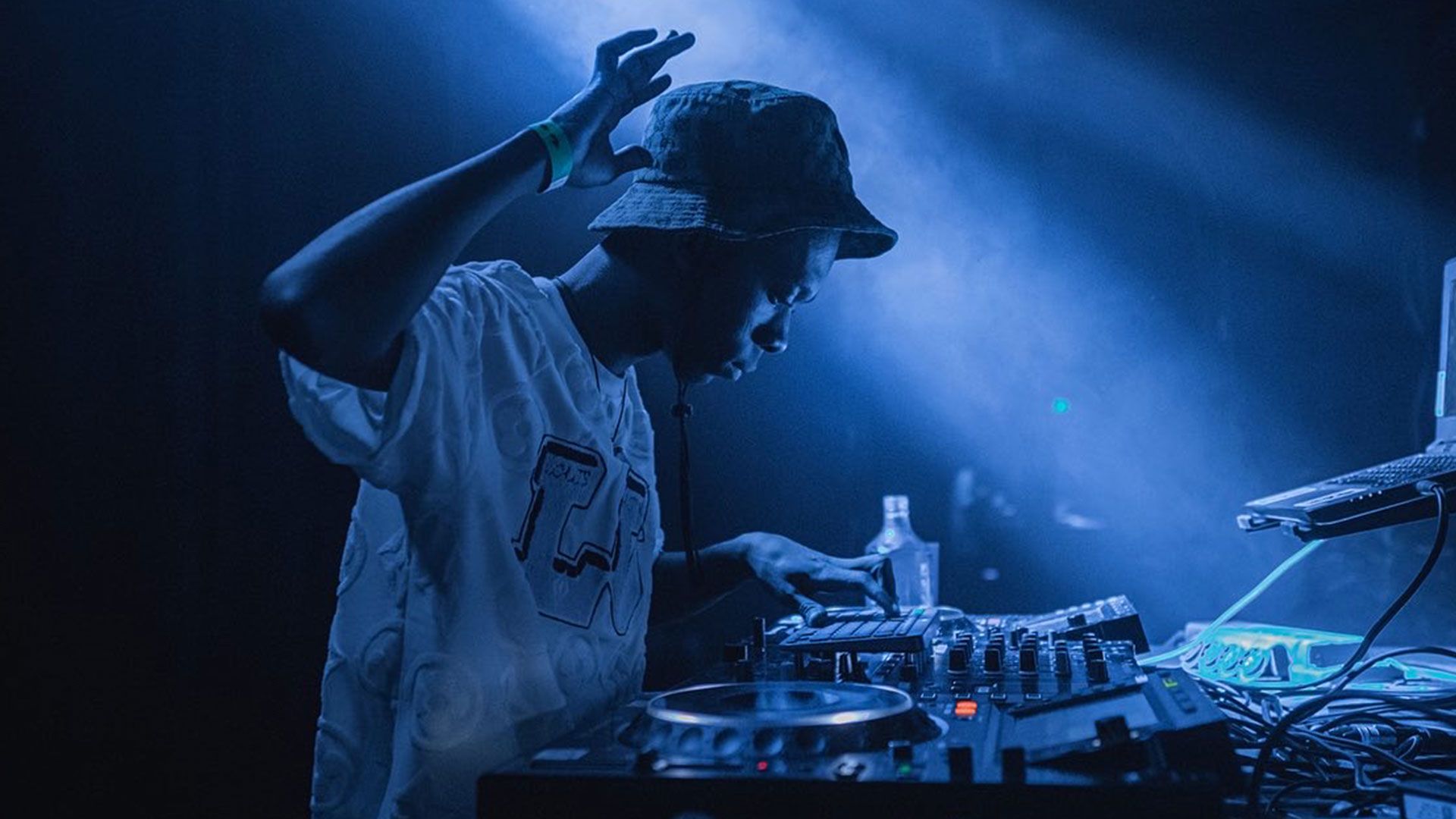 A side on view of DJ Travella behind DJ decks. One hand in the air.