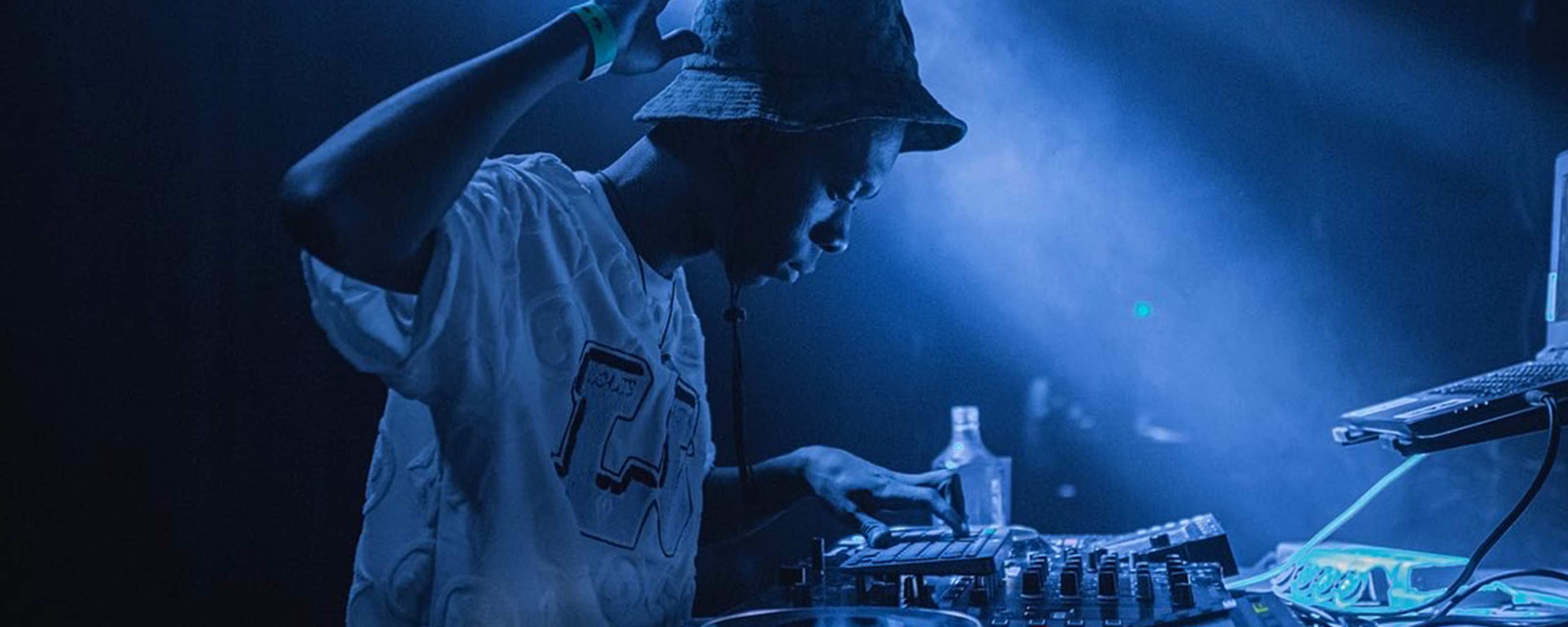 A side on view of DJ Travella behind DJ decks. One hand in the air.