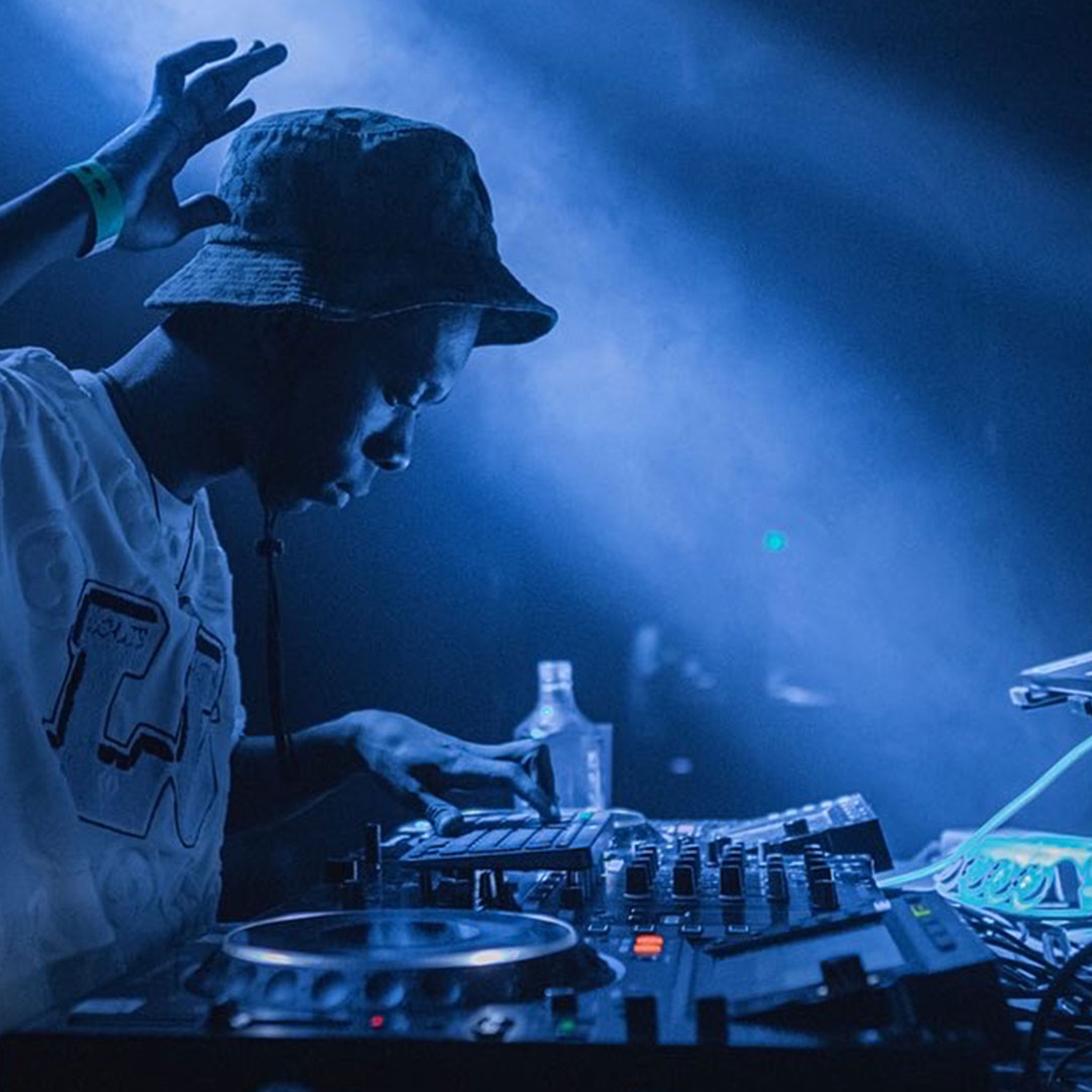 A DJ performs under moody lighting, concentrating on the decks with one hand raised mid-set.