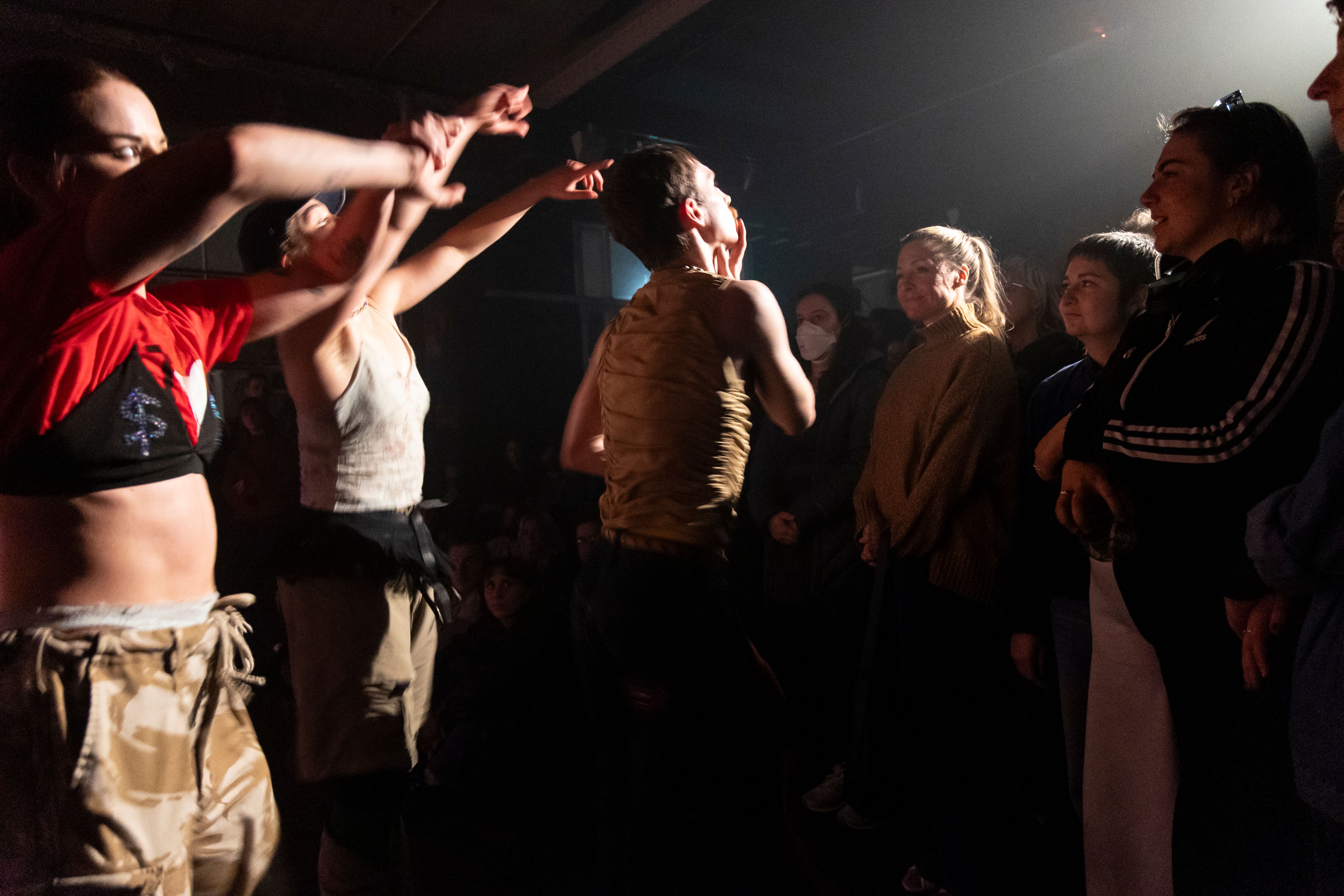 A trio of dancers perform close to a standing and sitting crowd, with a spotlight basking down on them from the back of the crowd.  