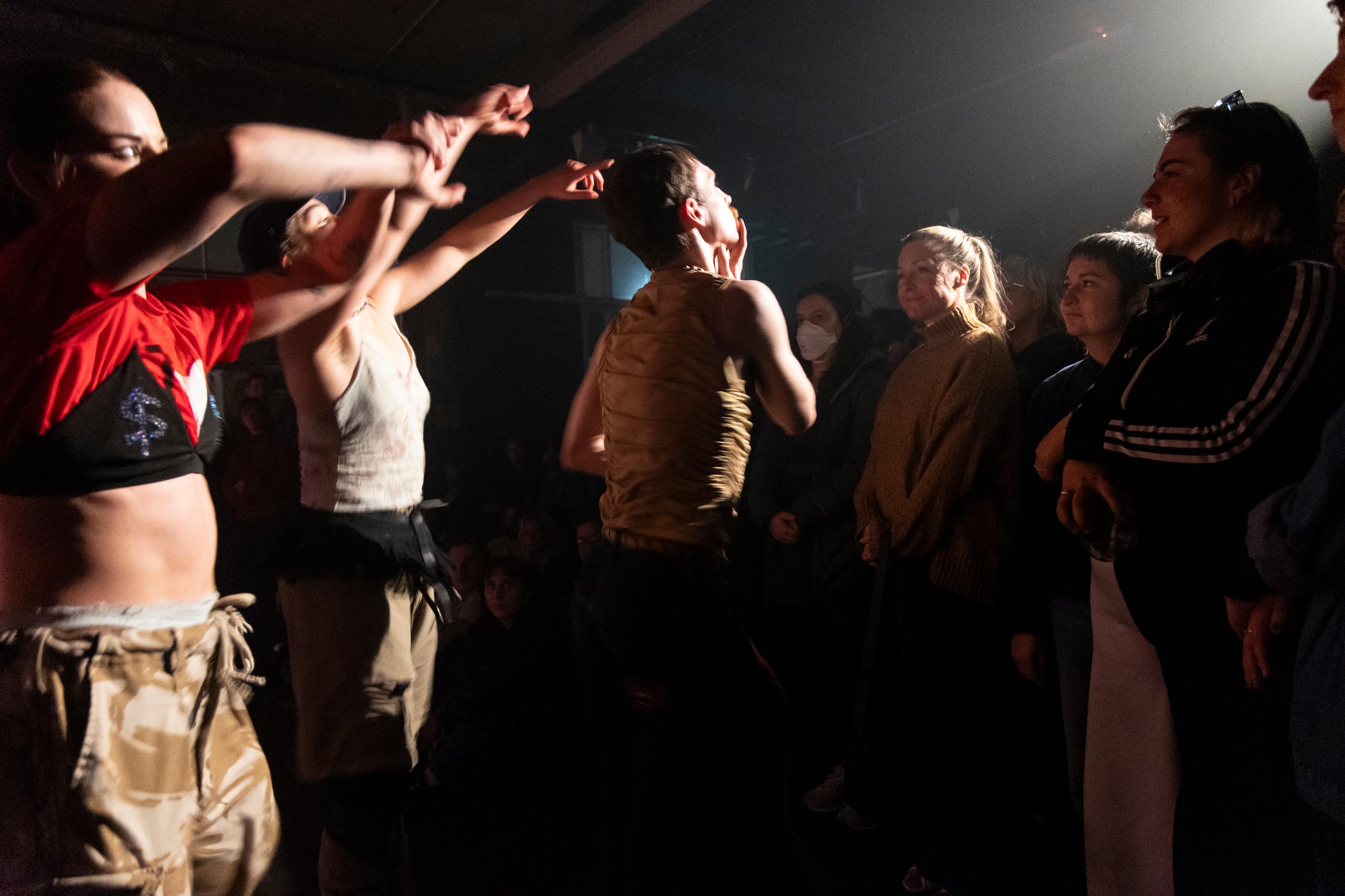 A trio of dancers perform close to a standing and sitting crowd, with a spotlight basking down on them from the back of the crowd.