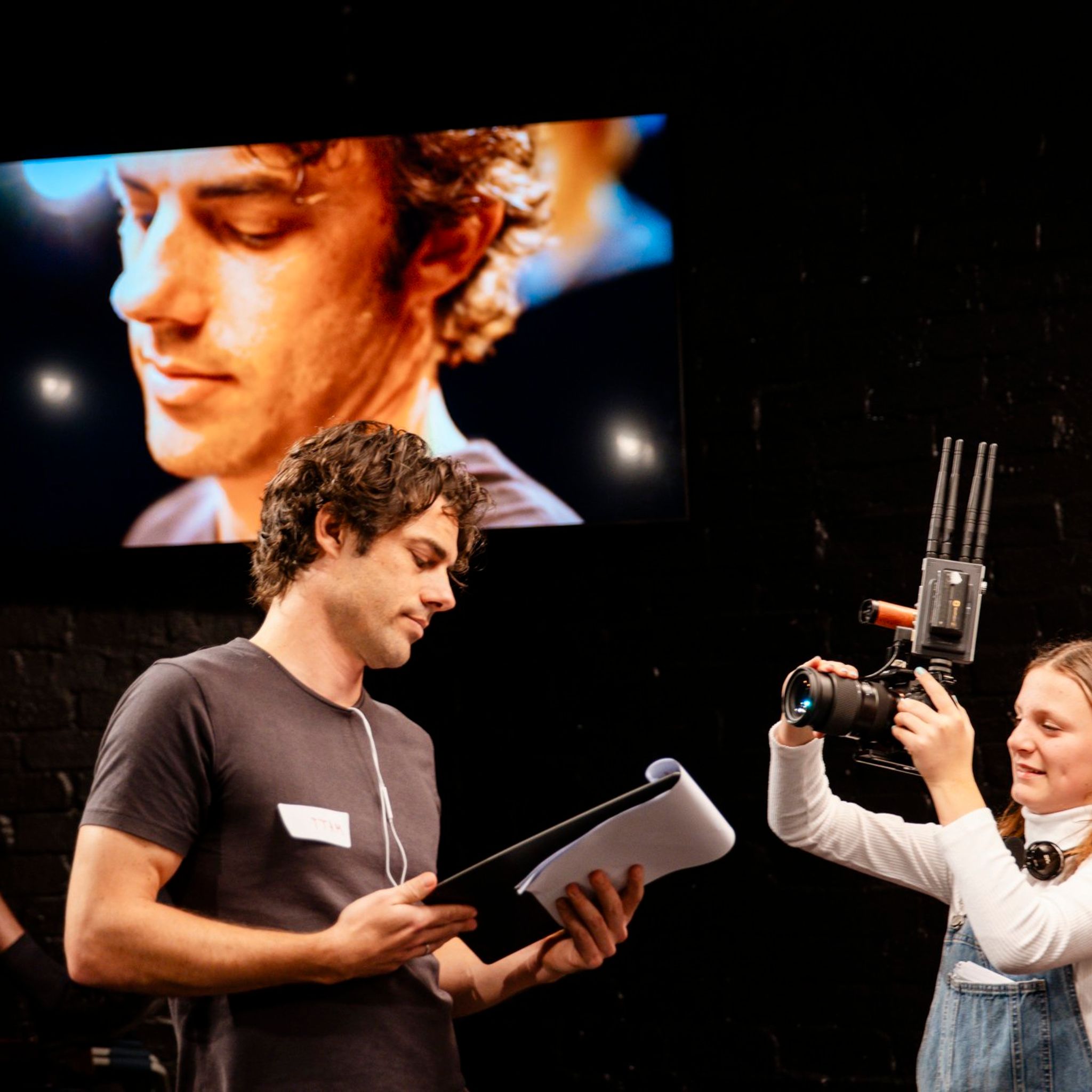 A young person holding a video camera films an adult man who is holding a script in his hand
