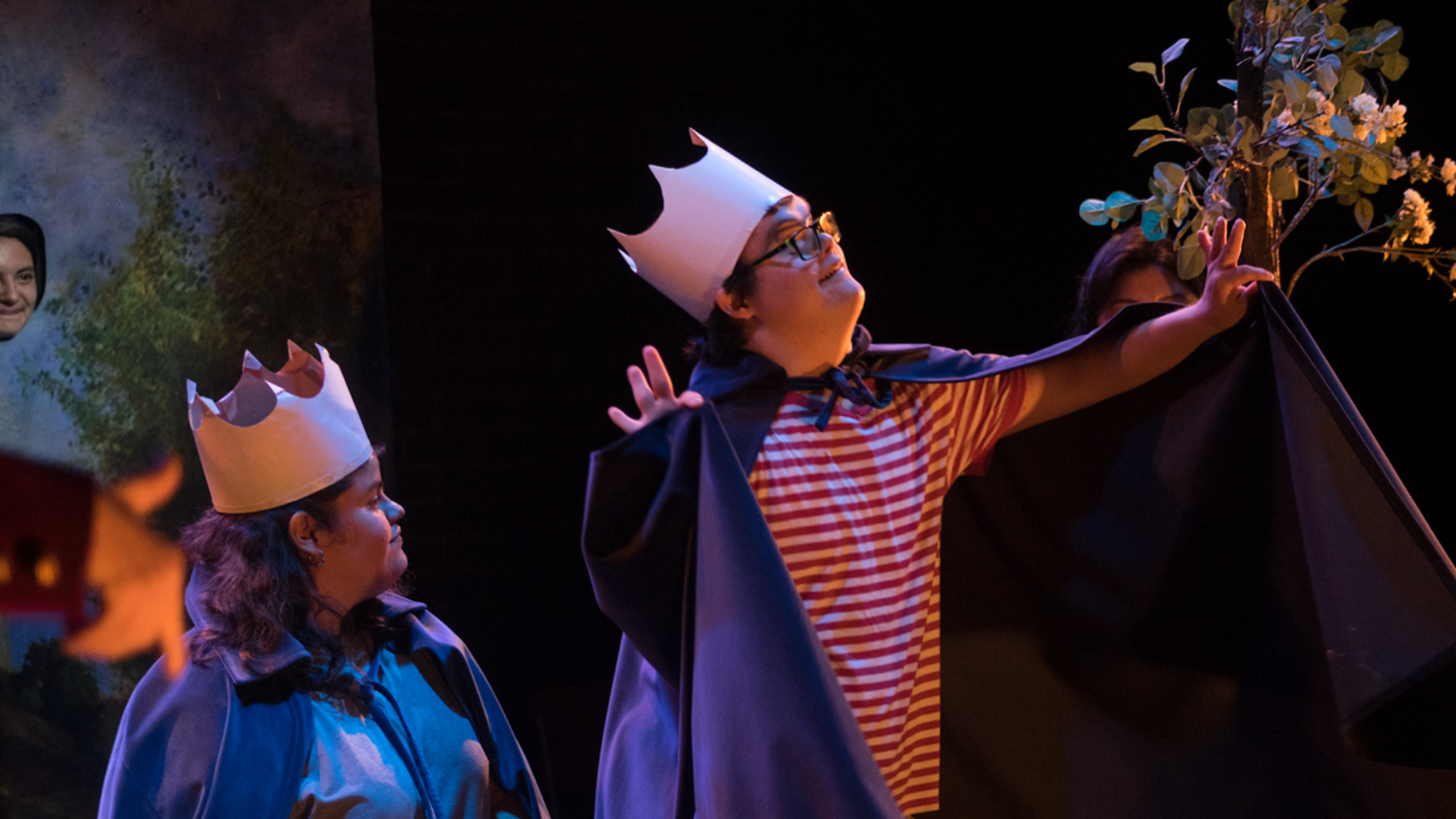 Two actors wearing capes and crowns gaze up towards a tree.