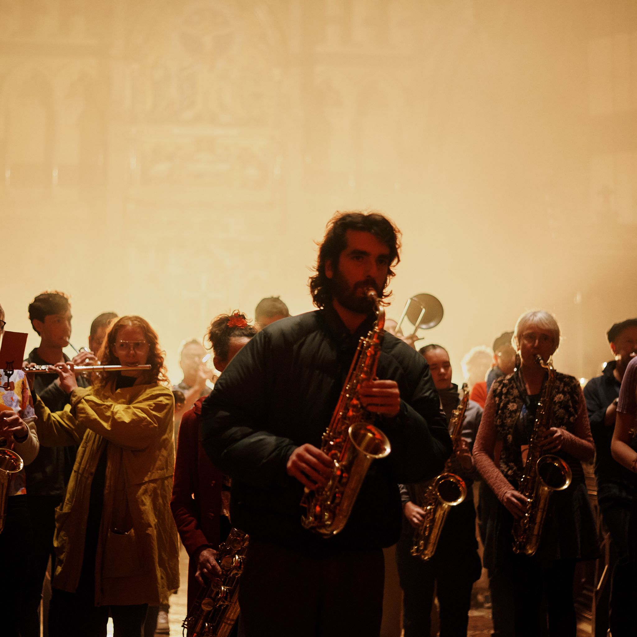 A crowd of musicians led by a saxophonist walk toward the camera, playing their instruments