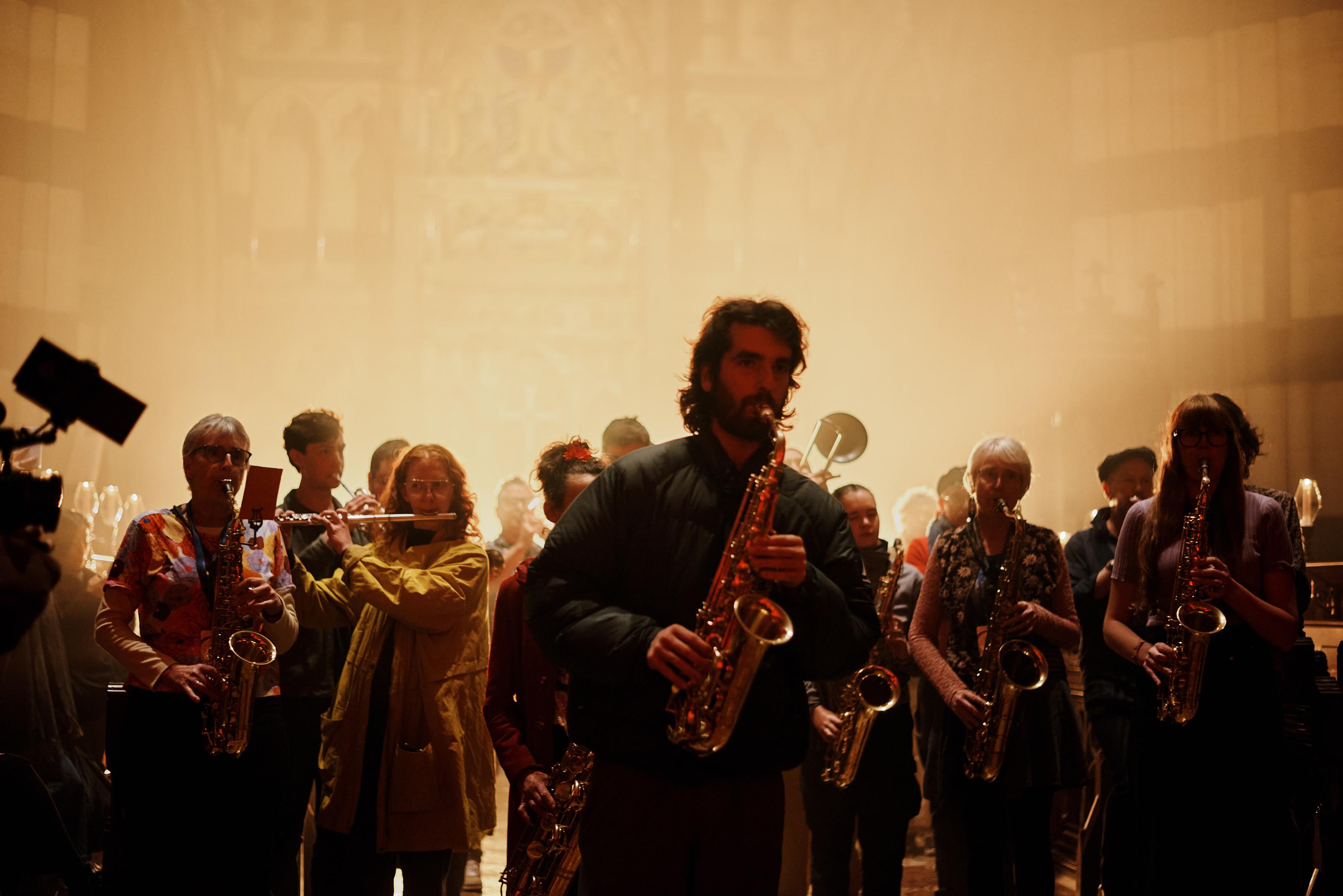 A crowd of musicians led by a saxophonist walk toward the camera, playing their instruments